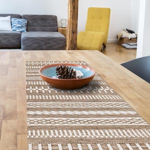 May include: A wooden dining table with a decorative woven table runner featuring a geometric pattern in brown and white. A ceramic bowl with a blue interior and brown exterior holds a pine cone and small white stones.
