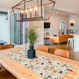 May include: A dining room table with a floral table runner. The table is made of light-colored wood and has black metal legs. The runner features a cream background with colorful flowers. Leather chairs surround the table, and a modern chandelier hangs above.