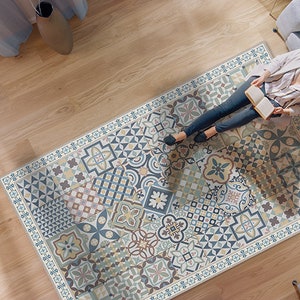 Puede incluir: Una alfombra estampada con una variedad de formas geométricas y colores, incluyendo azul, verde, amarillo y marrón. La alfombra está colocada sobre un suelo de madera y una persona está sentada en un sofá en el fondo.