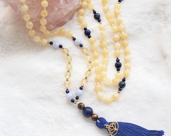 May include: A long beaded necklace with yellow, blue, and gold beads. The necklace has a blue tassel with a gold-coloured lotus flower charm. The beads are arranged in a layered design, with a large pink stone in the background.