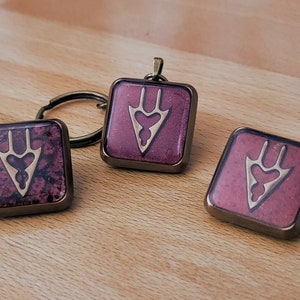 May include: Three square-shaped keyrings with a gold-coloured design of a stylised arrow with three points on a burgundy background. The keyrings are made of metal and have a shiny finish.