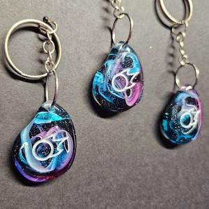 May include: Three resin keychains with silver key rings. Each keychain features a teardrop-shaped resin charm with a swirling blue, pink, and white design and a white symbol in the center.