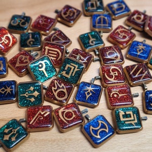 May include: A collection of 25 small, square, glittery charms with different symbols and designs. The charms are in shades of red, blue, and green, with gold accents. Each charm has a loop at the top for attaching to a necklace or bracelet.