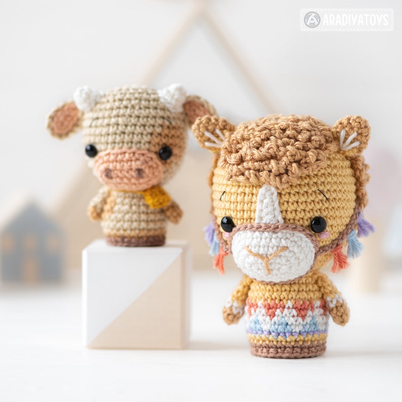 May include: Two crocheted stuffed toys, a brown and white cow and a yellow llama, both with black eyes and white noses. The cow is standing on a wooden block.