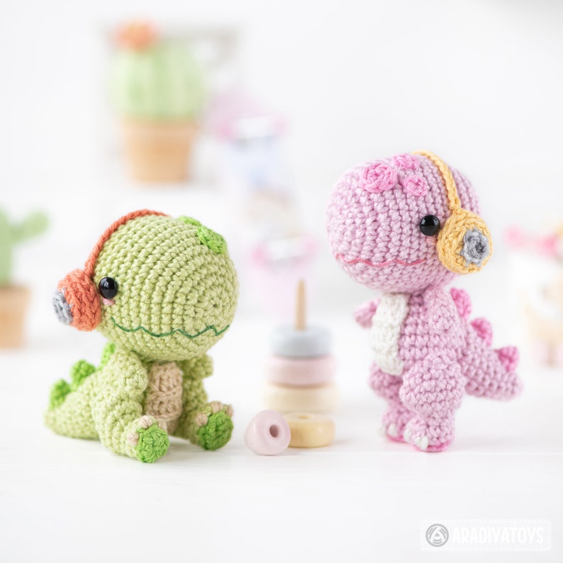 May include: Two crocheted dinosaur toys wearing headphones. The green dinosaur is sitting down and the pink dinosaur is standing up.