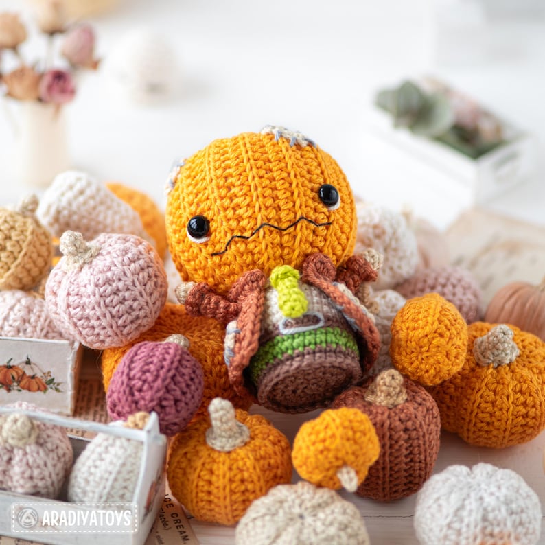 May include: A crocheted pumpkin character with black eyes and a stitched mouth, wearing a brown jacket, green shirt, and yellow tie. It is surrounded by numerous small, multi-colored crocheted pumpkins in shades of orange, pink, and brown.