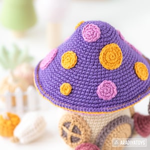 May include: A handmade crochet mushroom house with a purple cap adorned with orange and pink circular accents. The house features a tan base with a brown door and window. The whimsical design is perfect for children's decor.