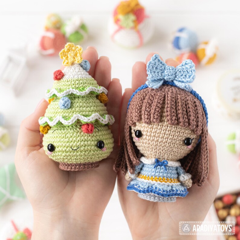 Op de afbeelding: Twee handgemaakte gehaakte figuren: een groene kerstboom met kleurrijke versieringen en een lachend gezicht, en een pop met bruin haar, een blauwe strik en een blauwe jurk. Andere gehaakte ornamenten staan op de achtergrond.