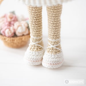 May include: Close-up of a crocheted doll's legs and feet. The legs are beige, and the feet are white, resembling ballet slippers. White yarn crisscrosses the ankles. A basket of pink and beige crocheted flowers is in the background.