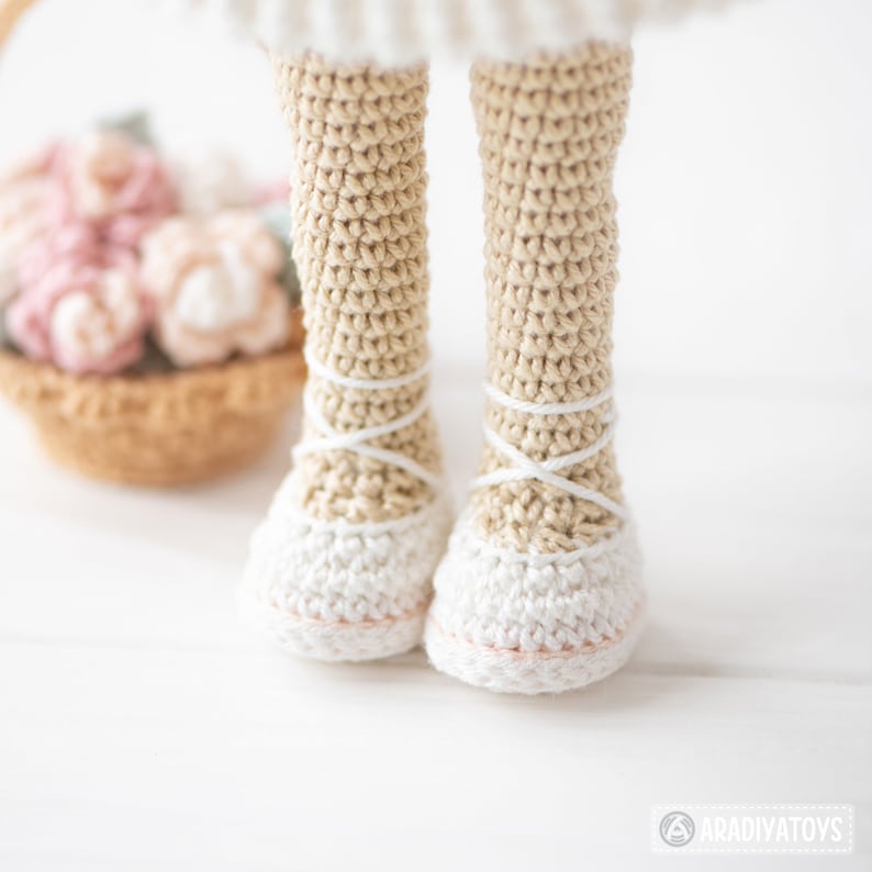May include: Close-up of a crocheted doll's legs and feet. The legs are beige, and the feet are white, resembling ballet slippers. White yarn crisscrosses the ankles. A basket of pink and beige crocheted flowers is in the background.