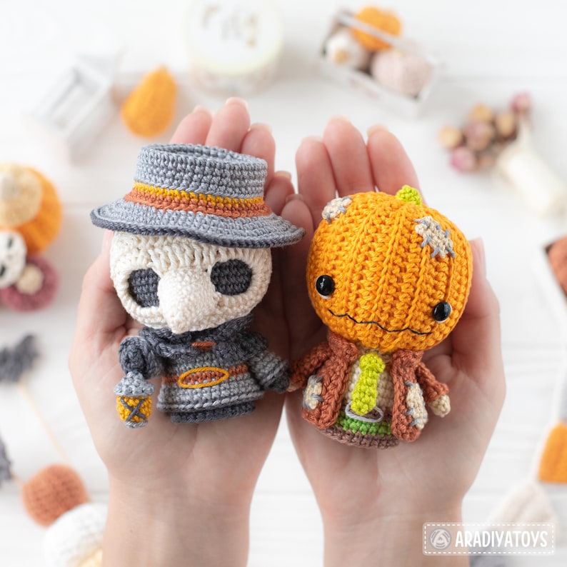 May include: Two crocheted dolls, one a plague doctor wearing a grey hat and coat with a white mask, and the other a pumpkin-headed character with a stitched orange head, brown jacket, and green tie. The text Aradiyatoys is visible in the corner.