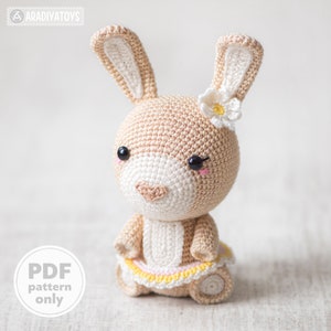 以下が含まれることがあります： 頭に白い花をつけた、かぎ針編みで作ったウサギのぬいぐるみです。黄色のスカートとピンクのスカートを着ています。ウサギは黒い目とピンクの鼻をしています。