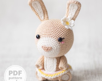 以下が含まれることがあります： 頭に白い花をつけた、かぎ針編みで作ったウサギのぬいぐるみです。黄色のスカートとピンクのスカートを着ています。ウサギは黒い目とピンクの鼻をしています。