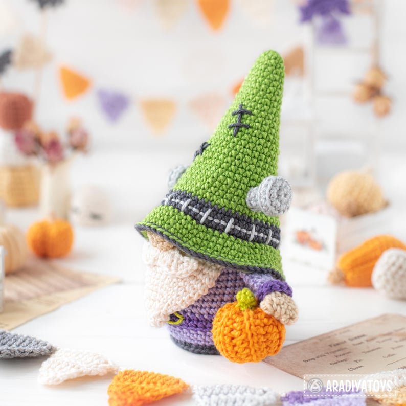 May include: A crocheted gnome doll featuring a tall, bright green pointed hat with dark grey stitching details and two light grey circular side bolts. The gnome has a cream-colored beard, a purple body, and is holding a small orange pumpkin.