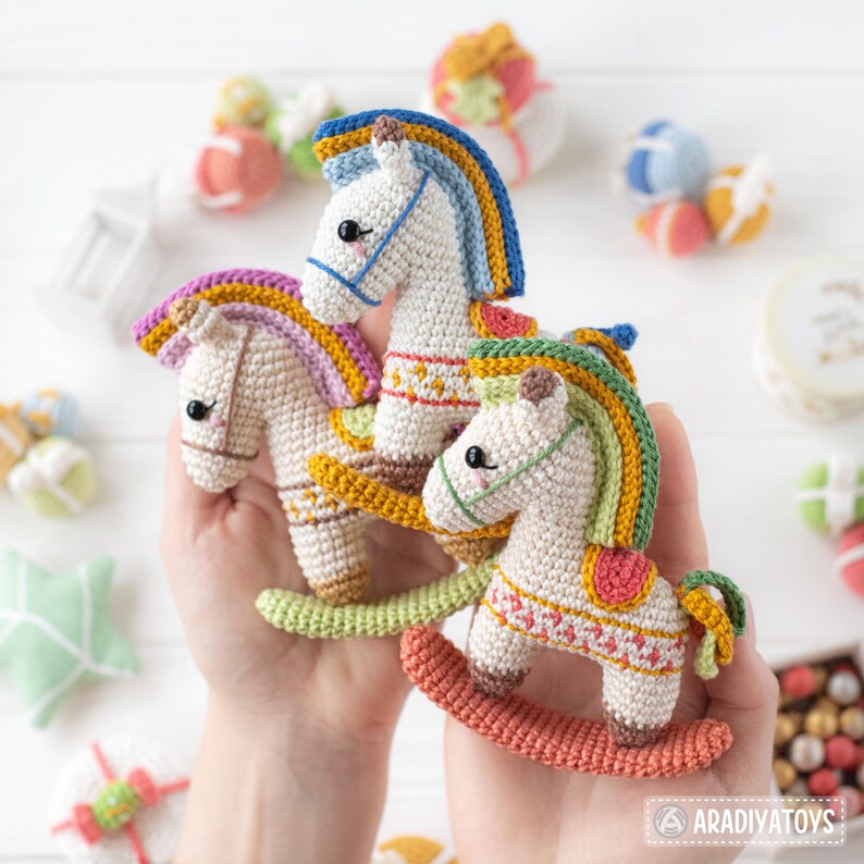 May include: Three handmade crochet rocking horses in cream, white, and tan. Each horse has a colorful mane and saddle, with details in blue, yellow, pink, and green. The horses are displayed against a white background with other small crochet toys.