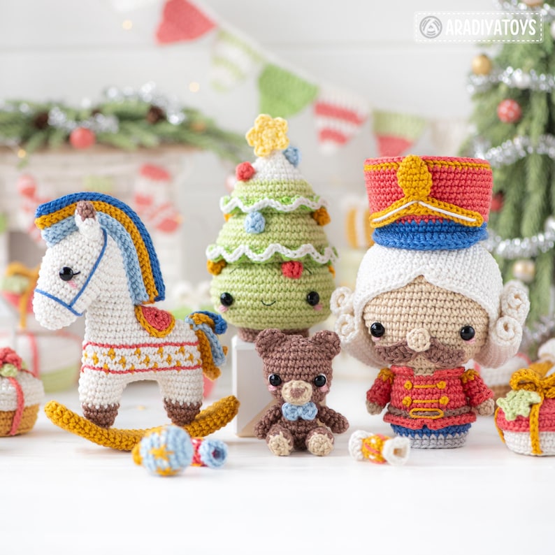 Op de afbeelding: Een verzameling handgemaakte gehaakte speelgoed, waaronder een wit schommelpaard met blauwe en gele accenten, een groene kerstboom, een bruine teddybeer met een blauwe strik en een rood-witte notenkraker. De speelgoed is op een witte ondergrond.