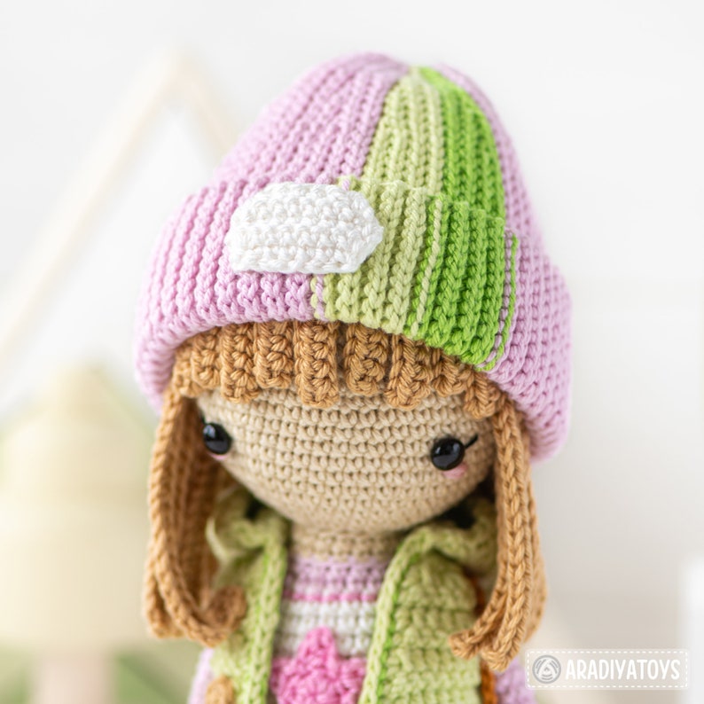 K&ouml;nnte beinhalten: H&auml;kelpuppe mit braunen Haaren, die einen rosa-gr&uuml;nen gestreiften Hut mit einem wei&szlig;en Patch tr&auml;gt. Die Puppe tr&auml;gt einen rosa-gr&uuml;nen gestreiften Pullover und ein rosa-wei&szlig; gestreiftes Hemd.