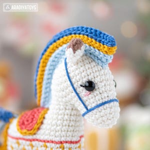 May include: A handmade crochet horse toy with a white body, blue, yellow, and light blue mane, and a pink and red saddle. The horse has black eyes and a blue bridle. The toy is a whimsical and colorful creation.