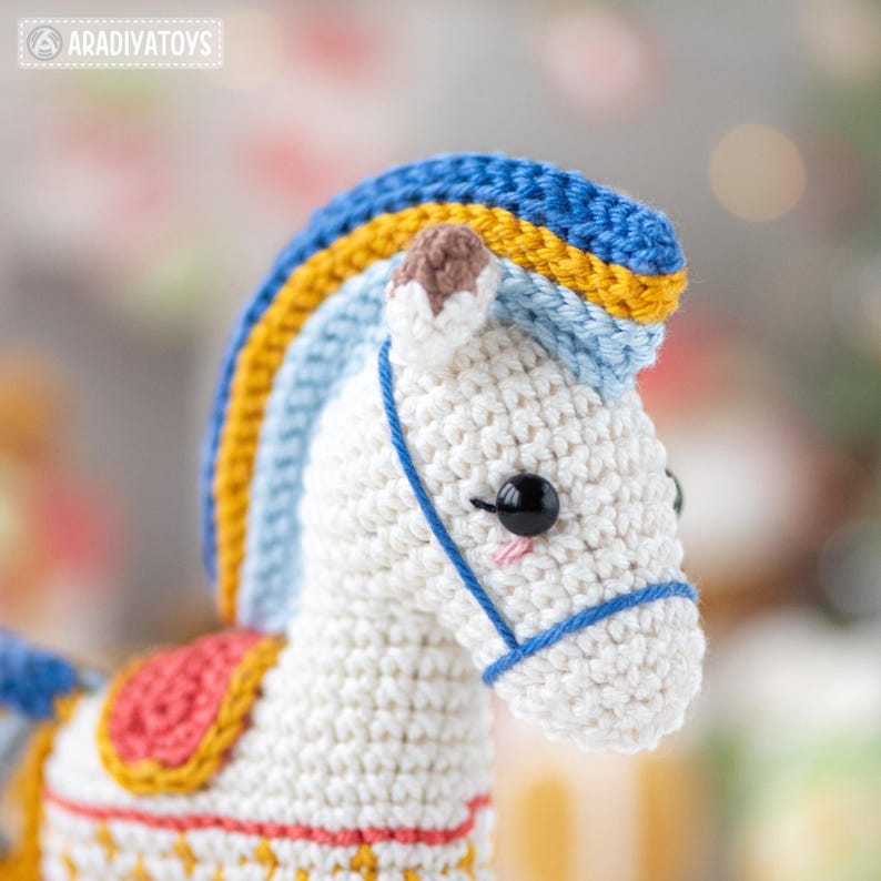 May include: A handmade crochet horse toy with a white body, blue, yellow, and light blue mane, and a pink and red saddle. The horse has black eyes and a blue bridle. The toy is a whimsical and colorful creation.