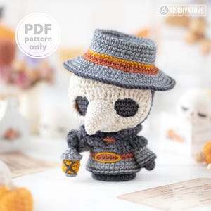 Può includere: Una bambola medico della peste all'uncinetto con un cappello grigio con fascia arancione, una maschera a becco color crema e un mantello grigio scuro con cintura arancione. La bambola tiene una piccola lanterna gialla. Testo: PDF pattern only.