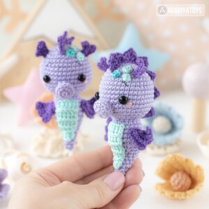 May include: Two handmade crochet seahorse toys in shades of purple and teal. The seahorses have black eyes and are displayed with other sea-themed decorations. The toys are approximately 4 inches tall.
