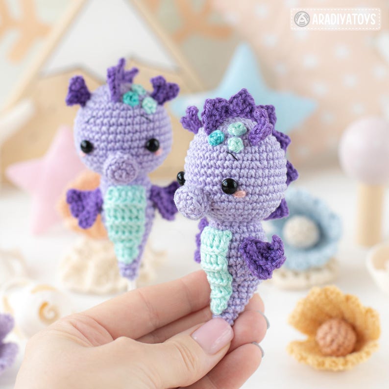 May include: Two handmade crochet seahorse toys in shades of purple and teal. The seahorses have black eyes and are displayed with other sea-themed decorations. The toys are approximately 4 inches tall.