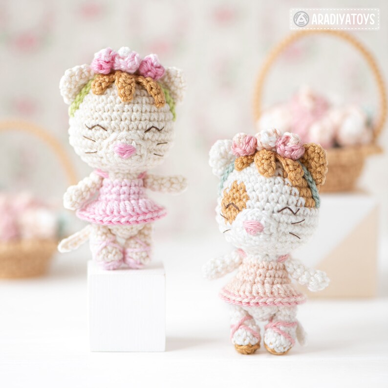 May include: Two handmade crochet cat dolls. The dolls are white with pink and brown accents, wearing pink dresses and flower crowns. One doll is standing on a white block. The background includes baskets and a floral pattern.