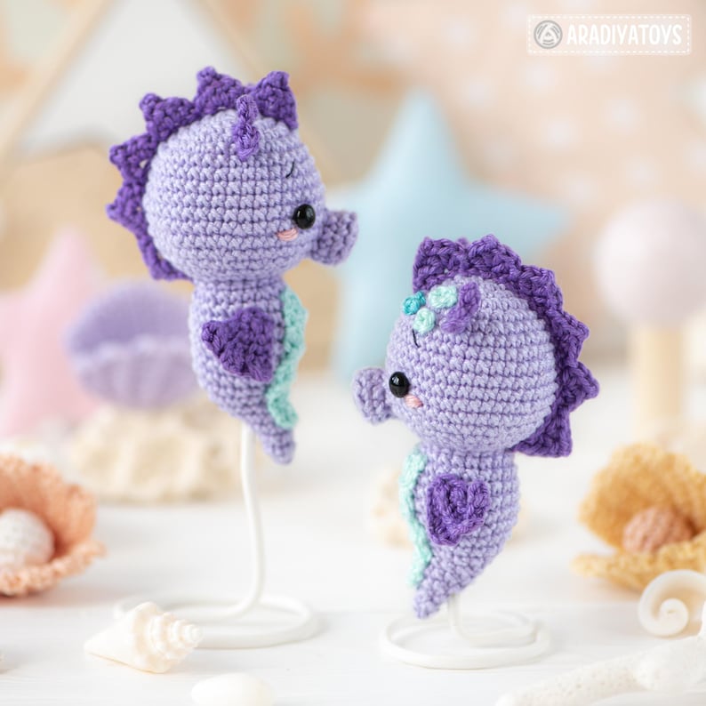 May include: Two handmade crochet seahorse figurines in shades of purple and teal. The seahorses have a heart-shaped detail and are mounted on white stands. The background includes seashell decorations and a star-shaped ornament.