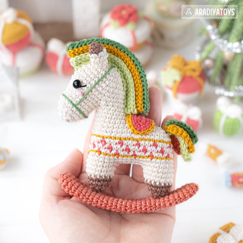 May include: A handmade crochet rocking horse in cream, with a rainbow mane and tail. The horse has a pink and yellow saddle and a red and yellow patterned design on its body. The rocking horse is a charming, handcrafted toy.
