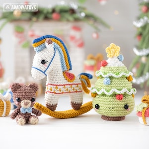 Op de afbeelding: Een verzameling handgemaakte gehaakte kerstversieringen. De set bevat een wit schommelpaard met een blauwe en gele manen, een bruine teddybeer met een blauwe strik en een groene kerstboom met kleurrijke ornamenten en een gele ster.