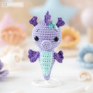 May include: A handmade crochet seahorse toy in shades of purple and seafoam green. The seahorse has black safety eyes and a small flower detail on its head. The toy is displayed on a white stand, with other crochet sea-themed items in the background.