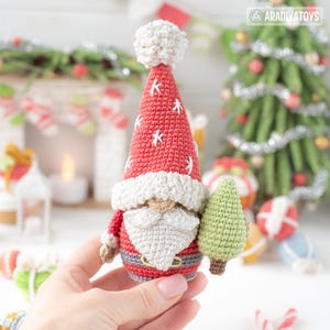 K&ouml;nnte beinhalten: Handgefertigte geh&auml;kelte Weihnachtsmann-Figur mit roter M&uuml;tze und Mantel, wei&szlig;em Bart und grauem G&uuml;rtel. Der Hut ist mit wei&szlig;en Sternen verziert. Der Weihnachtsmann h&auml;lt einen kleinen gr&uuml;nen geh&auml;kelten Weihnachtsbaum. Im Hintergrund befinden sich Weihnachtsbaum und Dekorationen.