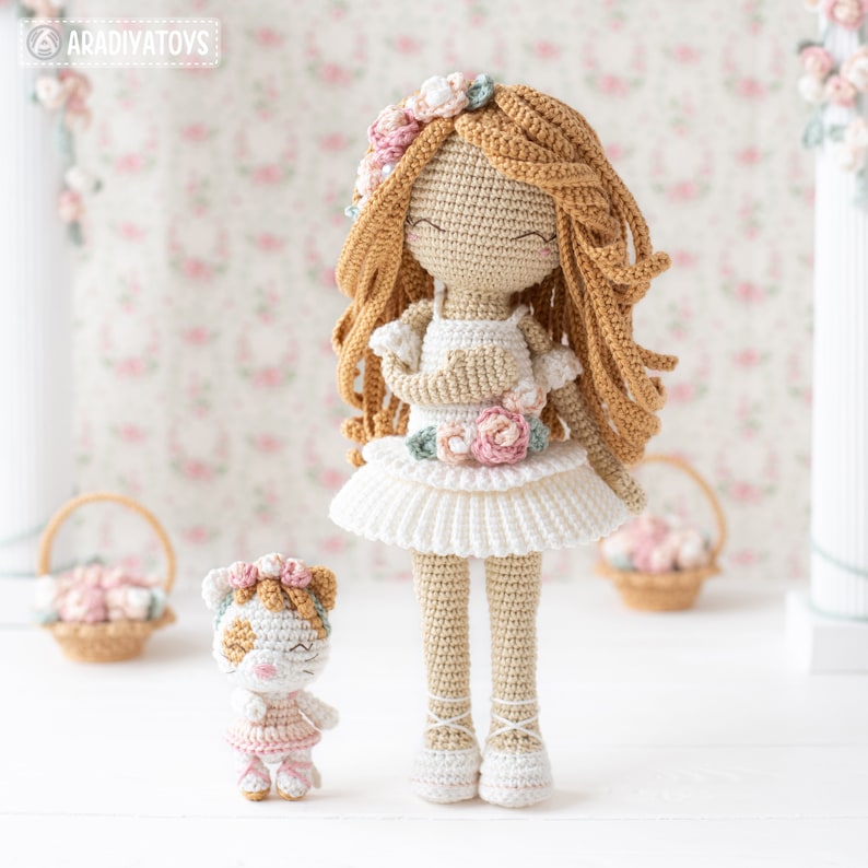 May include: A pair of handmade crochet dolls. The larger doll has long, flowing, light brown hair and wears a white dress with floral accents. The smaller doll, resembling a cat, is in a pink dress. Both dolls have floral headbands and are on a white surface.