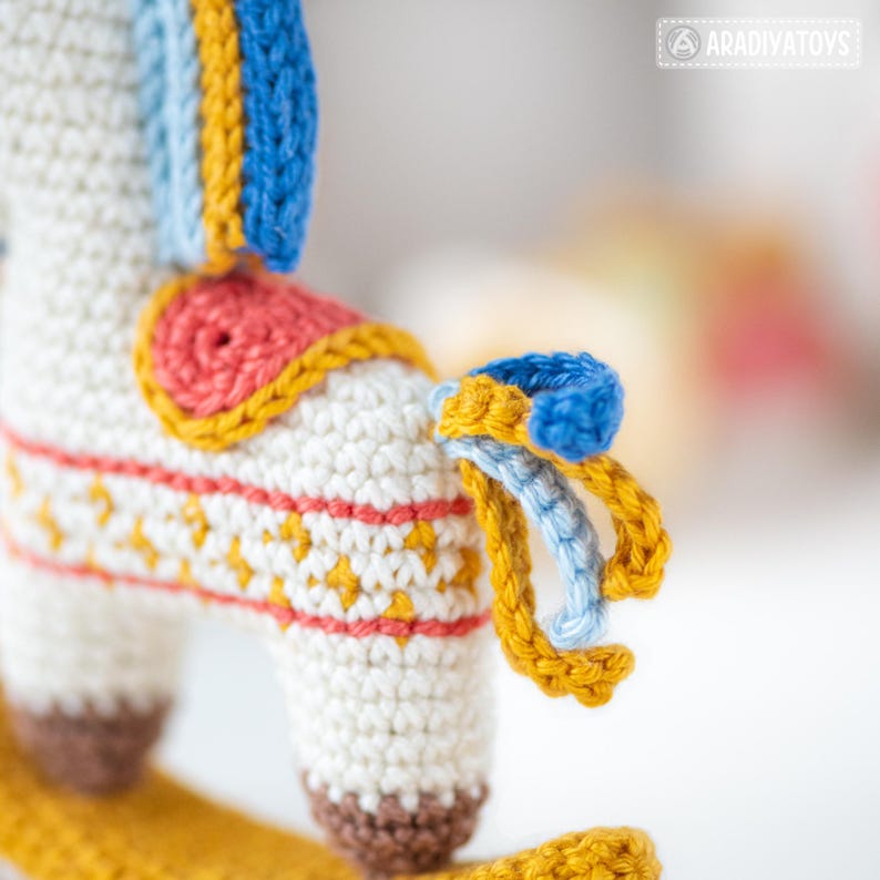 May include: Close-up of a handmade crochet rocking horse toy. The toy features a white body with colorful accents, including blue, yellow, and red details. The horse has a detailed saddle and mane, and a yellow base. The toy is designed for children.