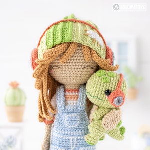 May include: A crocheted doll with long brown hair wearing a green and orange striped hat and blue overalls. The doll is holding a green crocheted dinosaur with orange headphones.