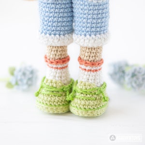 May include: Crocheted doll wearing blue jeans, white socks with orange stripes, and green shoes. The doll is standing on a white surface.