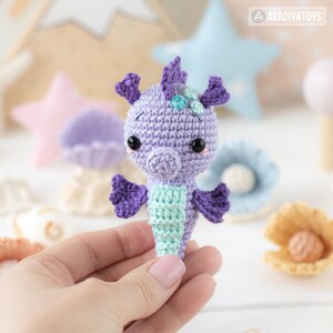 May include: A handmade crochet seahorse in shades of purple and teal. The seahorse has black eyes and small fins. The background includes star-shaped decorations and a seashell. The seahorse is held in a person's hand.