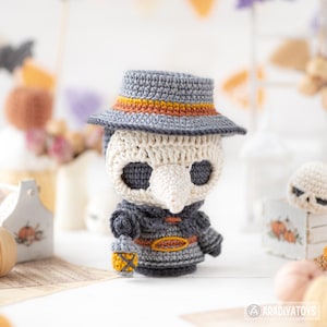 May include: A crocheted plague doctor doll featuring a cream-colored bird-like mask, dark gray eye sockets, and a gray hat with an orange band. The figure wears a dark gray coat with an orange belt and holds a small orange lantern in one hand.