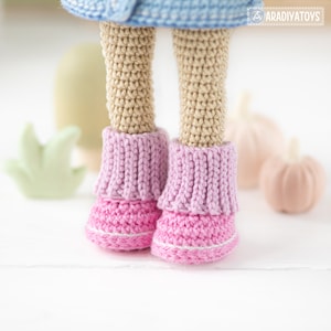 K&ouml;nnte beinhalten: Nahaufnahme der Beine einer geh&auml;kelten Puppe, die rosa und lila Stiefel tr&auml;gt. Die Puppe steht auf einer wei&szlig;en Oberfl&auml;che mit ein paar kleinen geh&auml;kelten K&uuml;rbissen im Hintergrund.