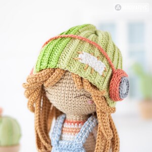 May include: A crocheted doll with long brown hair wearing a green and white striped hat with a red and gray earbud. The doll is wearing a blue and white striped shirt and blue overalls.