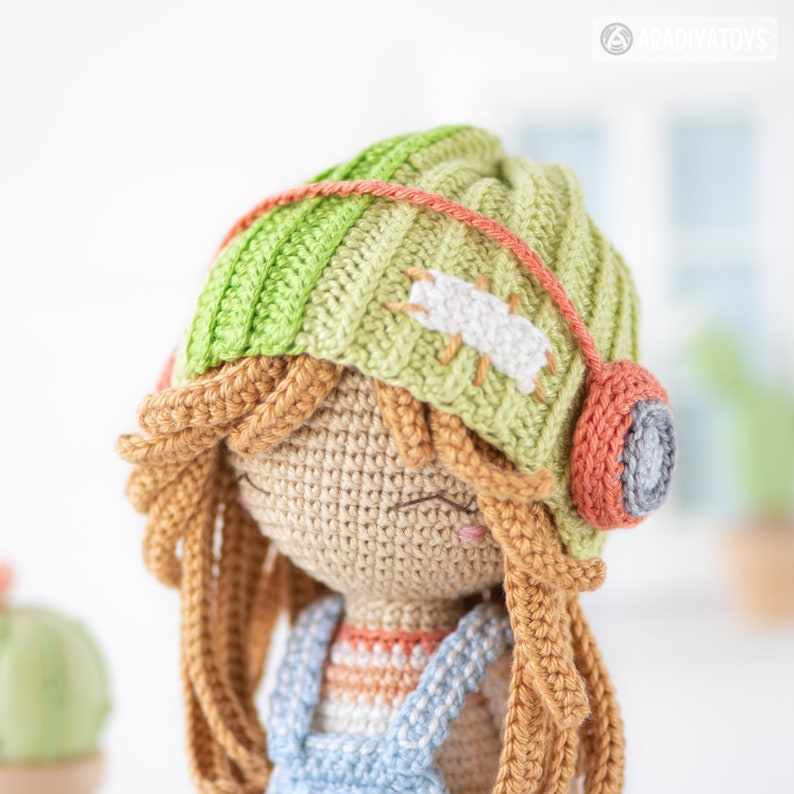 May include: A crocheted doll with long brown hair wearing a green and white striped hat with a red and gray earbud. The doll is wearing a blue and white striped shirt and blue overalls.
