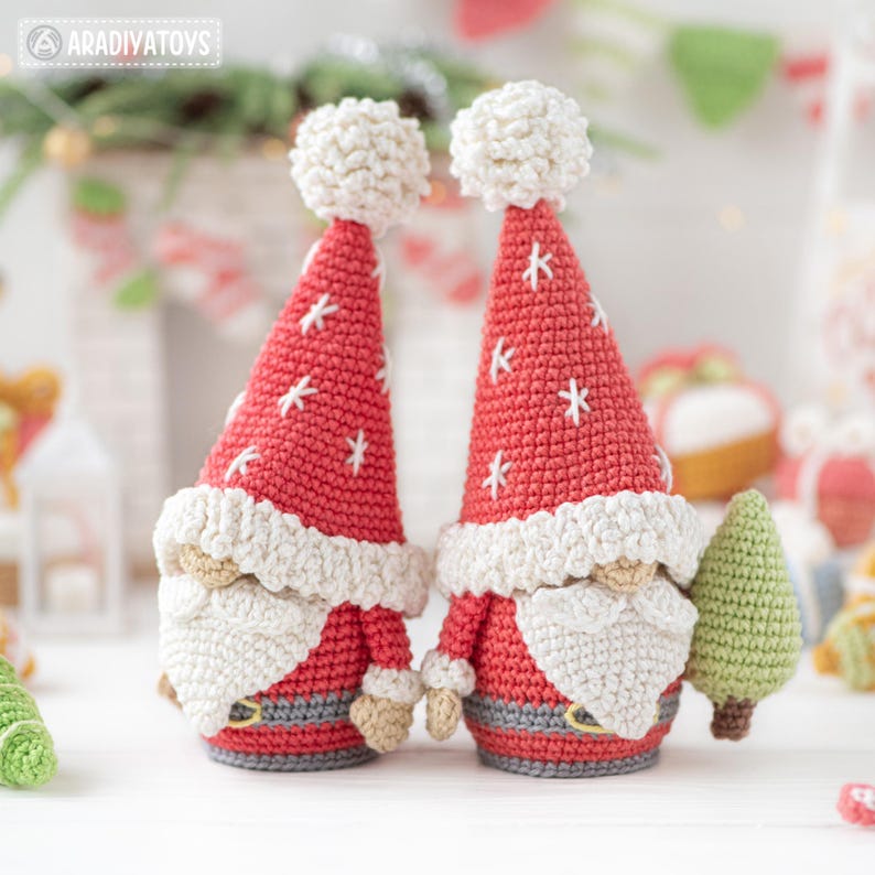 K&ouml;nnte beinhalten: Zwei handgefertigte geh&auml;kelte Weihnachtsmann-Zwerge. Jeder Zwerg hat eine rote M&uuml;tze mit wei&szlig;en Schneeflocken-Akzenten und einem wei&szlig;en Bommel. Sie haben wei&szlig;e B&auml;rte, graue G&uuml;rtel und halten einen kleinen gr&uuml;nen Weihnachtsbaum. Der Hintergrund ist unscharf.
