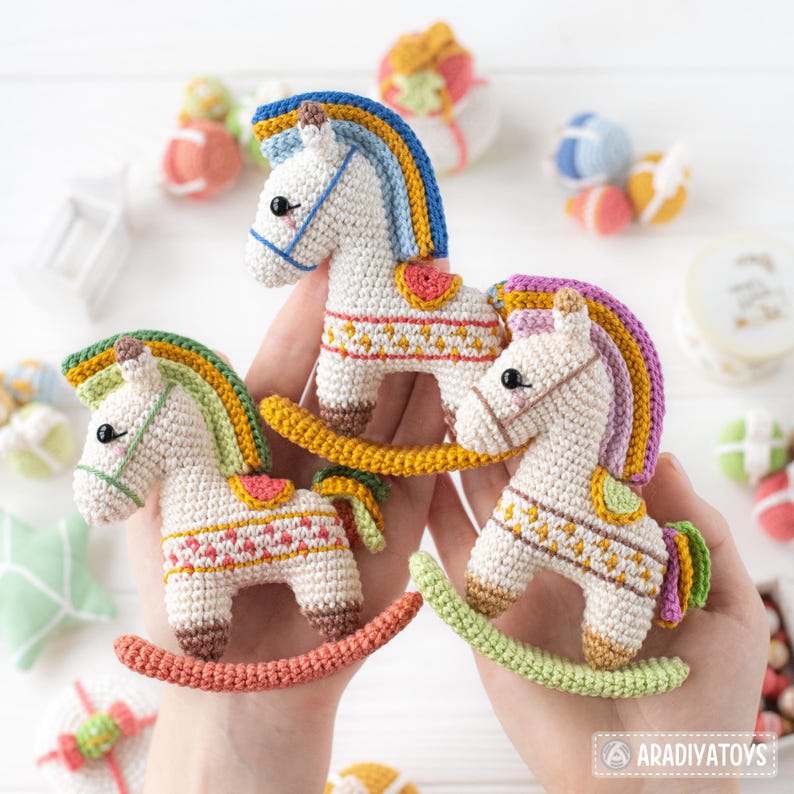 May include: Three handmade crochet rocking horse toys in white, with colorful manes and saddles. Each horse features a unique color scheme, including blue, green, and purple manes, and is held in a person's hands. The toys are decorated with intricate patterns.