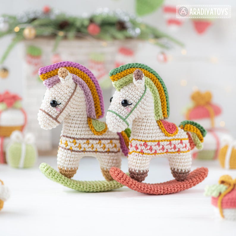 May include: Two handmade crochet rocking horses. The horses are white with colorful manes, saddles, and decorative patterns. One horse has a green rocker, and the other has a coral rocker. The background includes Christmas decorations and gift boxes.