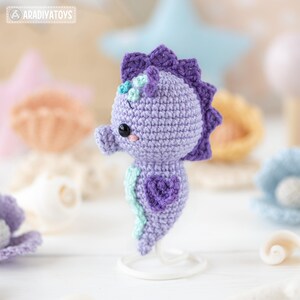 May include: A handcrafted, crochet seahorse in shades of purple, with a heart-shaped design on its body. The seahorse has a decorative fin and a small flower on its head. The image includes the text "ARADIYATOYS".