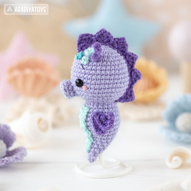 May include: A handcrafted, crochet seahorse in shades of purple, with a heart-shaped design on its body. The seahorse has a decorative fin and a small flower on its head. The image includes the text "ARADIYATOYS".