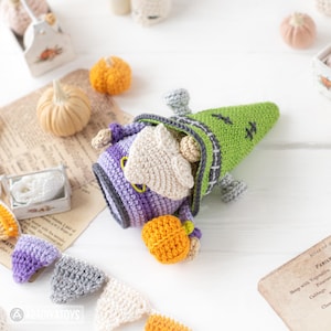 May include: A crocheted gnome figure featuring a green pointed hat with black stitch details, a purple body, and a beige beard. The gnome holds a small orange crocheted pumpkin in its hands. A crocheted bunting banner with purple, gray, and white triangles is positioned below the figure.