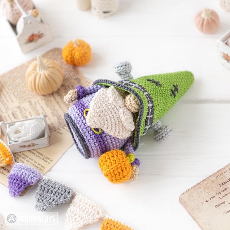 May include: A crocheted gnome figure featuring a green pointed hat with black stitch details, a purple body, and a beige beard. The gnome holds a small orange crocheted pumpkin in its hands. A crocheted bunting banner with purple, gray, and white triangles is positioned below the figure.