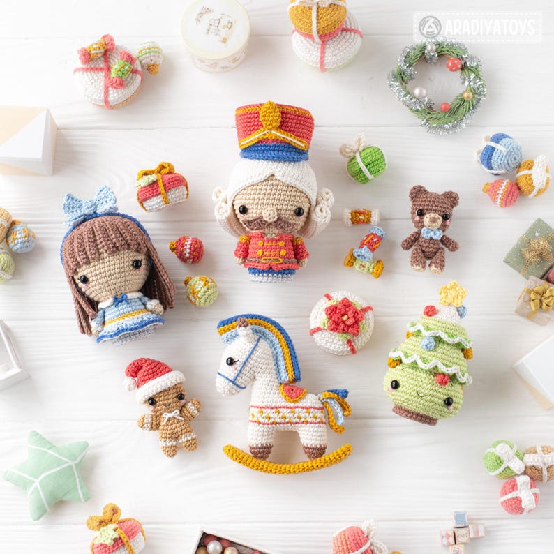 Op de afbeelding: Een verzameling handgemaakte gehaakte speelgoed, waaronder een notenkraker, een pop, een schommelpaard, een teddybeer en een kerstboom. Het speelgoed is in verschillende kleuren, zoals rood, blauw, groen en wit. Het speelgoed is op een wit oppervlak geplaatst.