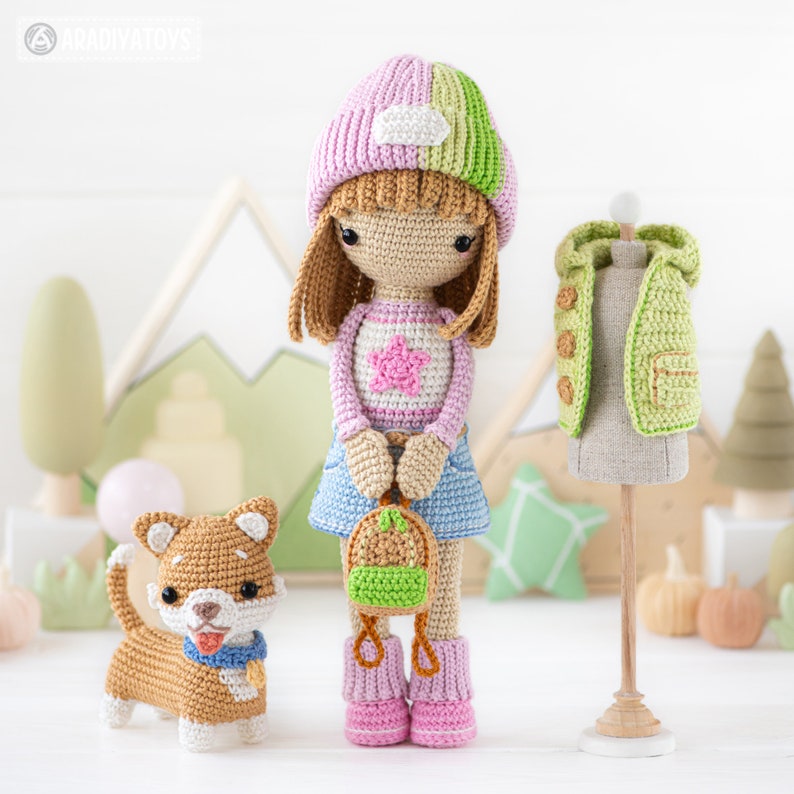 K&ouml;nnte beinhalten: Eine geh&auml;kelte Puppe mit einem rosa-gr&uuml;nen Hut, einem rosa-wei&szlig; gestreiften Shirt mit einem rosa Stern, einem blauen Jeansrock und rosa Stiefeln. Die Puppe h&auml;lt einen gr&uuml;n-braunen Rucksack. Ein brauner geh&auml;kelter Hund mit einem blauen Halsband steht neben der Puppe. Eine gr&uuml;ne geh&auml;kelte Weste steht auf einem Holzst&auml;nder hinter der Puppe.